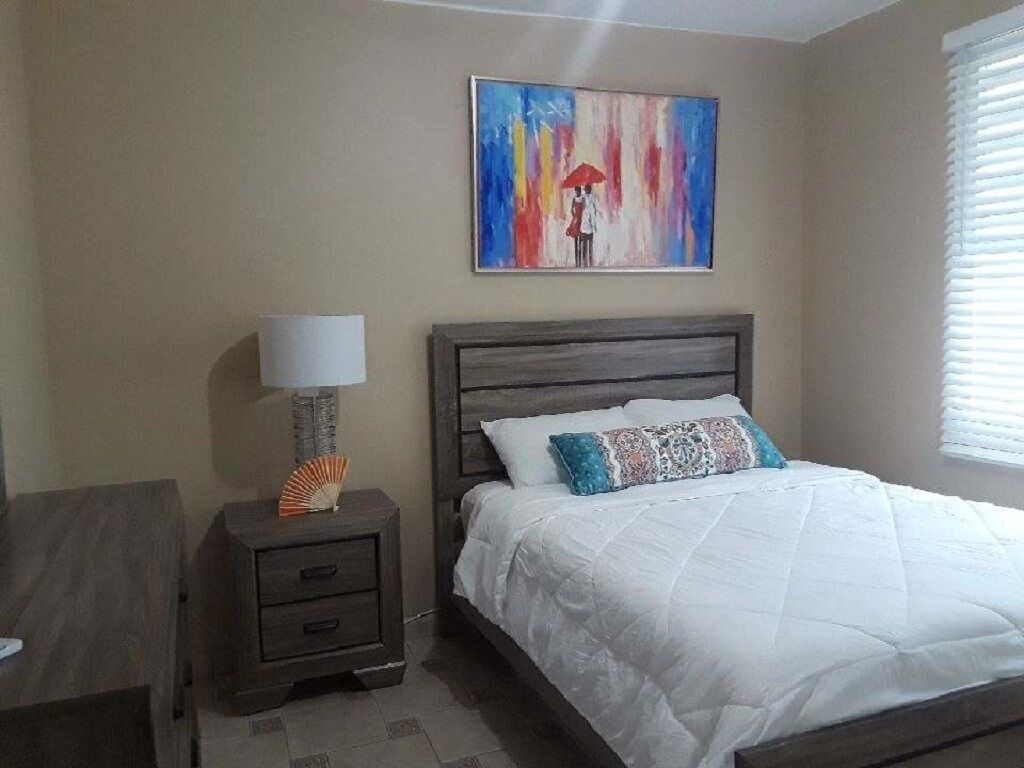 A bedroom with a bed, a nightstand with a lamp, a painting on the wall, and a window with blinds.