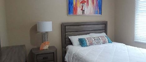 A bedroom with a bed, a nightstand with a lamp, a painting on the wall, and a window with blinds.