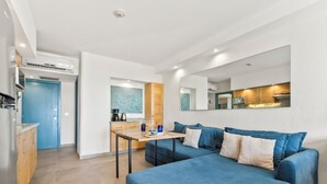Een knus kamertje met een blauwe bank, een klein houten tafeltje en een kitchenette.