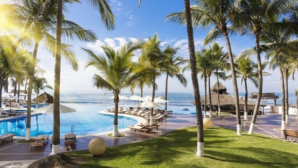 Zwembadgedeelte van een resort met palmbomen, ligbedden en uitzicht op de oceaan.