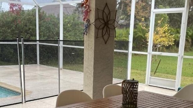 A covered patio with a table and chairs, a decorative wall piece, and a view of a swimming pool and greenery. - Quiet, Cozy, Bungalow with a Pool. Near Venice Beach and Warm Mineral Springs (North Port)