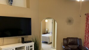 A living room with a flat-screen TV, a leather recliner, a white entertainment unit, a ceiling fan, and a hallway leading to a bedroom. - Quiet, Cozy, Bungalow with a Pool. Near Venice Beach and Warm Mineral Springs (North Port)