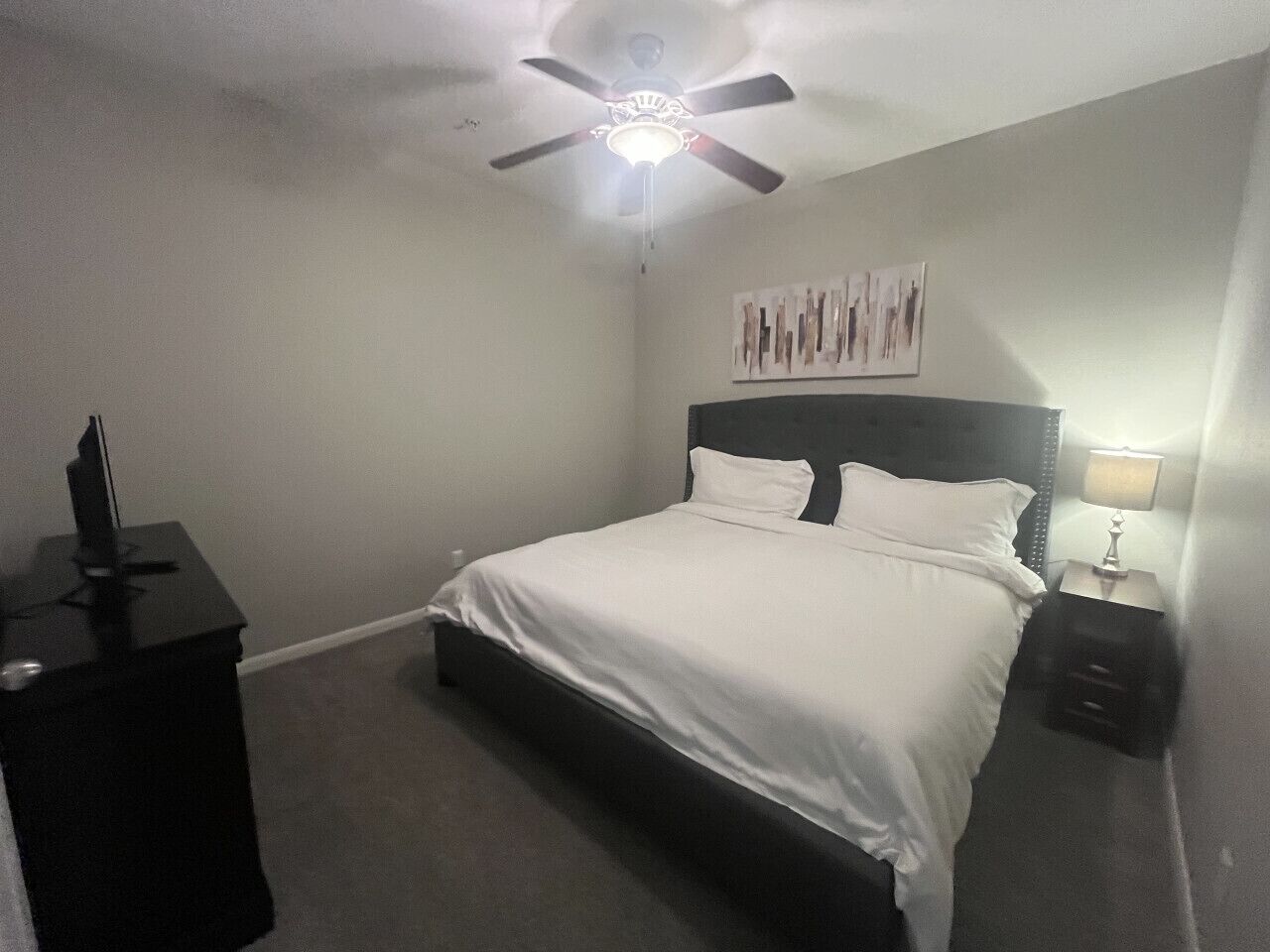 A bedroom with a bed, a ceiling fan, a nightstand, and a wall-mounted artwork.
