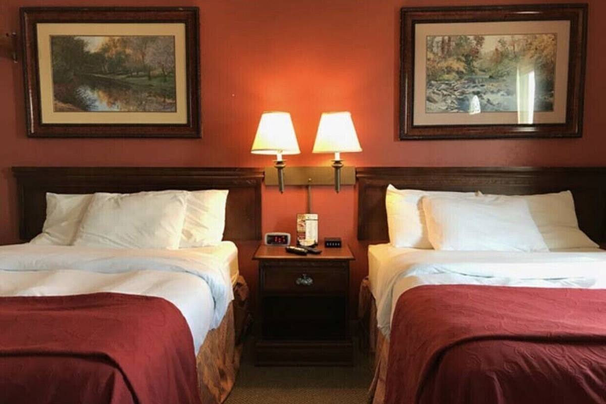 Quarto de hotel com duas camas, cada uma com roupa de cama branca, uma mesa de cabeceira com relógio e abajur, e duas fotografias emolduradas nas paredes.