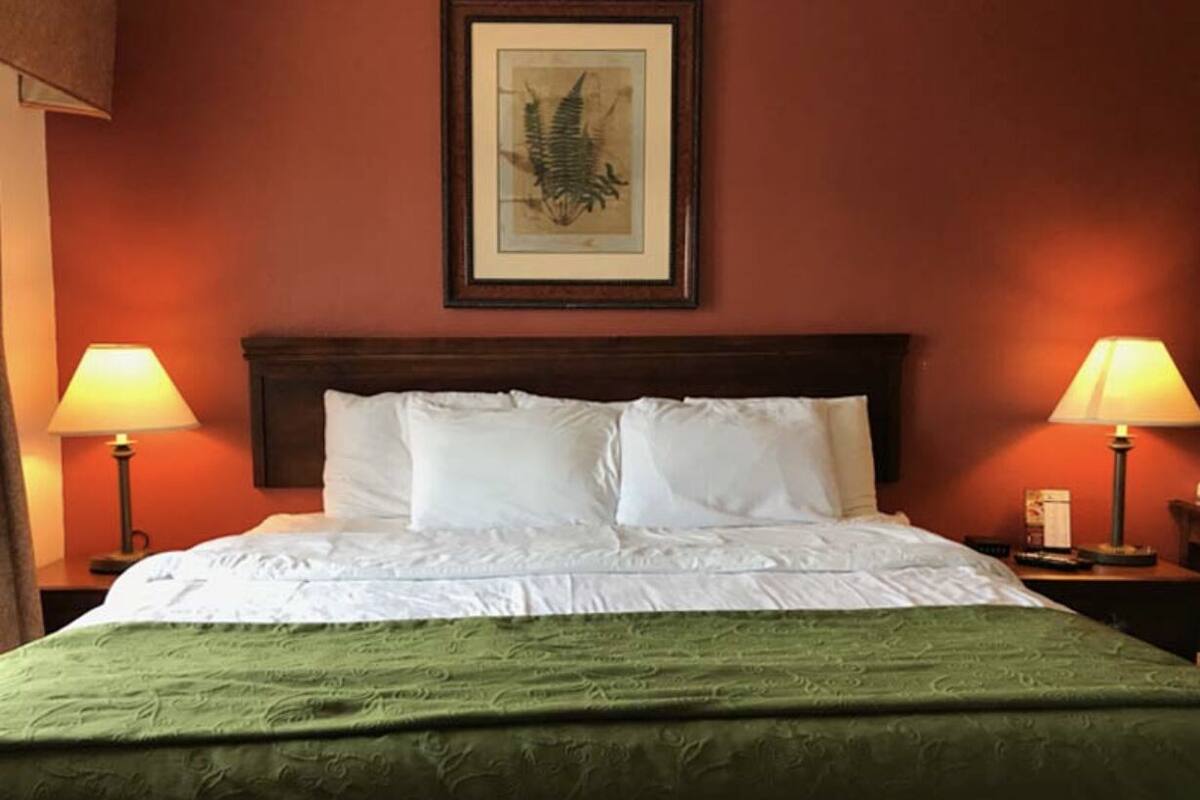 Cama bem arrumada com lençóis brancos e manta verde, com duas luminárias de cabeceira em mesas noturnas idênticas.