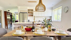 Un comedor moderno con una mesa de madera dispuesta para seis personas, rodeada de sillas grises tapizadas. Hay un tazón rojo grande lleno de fruta y una botella de vino sobre la mesa.