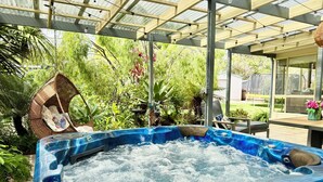 Un jacuzzi bajo un porche cubierto con estructura de madera y luces colgantes, rodeado de exuberante vegetación.