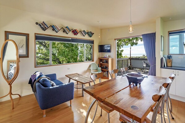 A modern living room with a wooden dining table, blue sofa, and a large window offering a view of greenery.