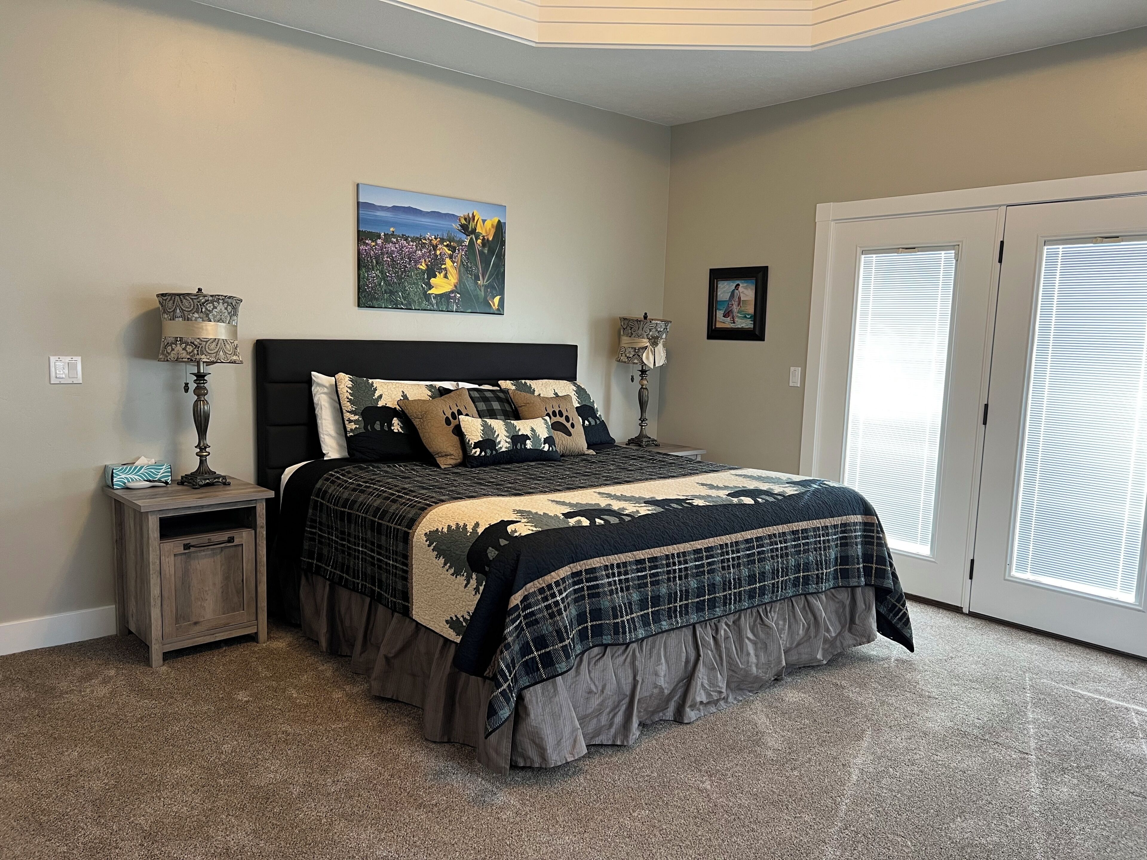 A bedroom with a large bed, two bedside tables, a nightstand, a window with blinds, and a painting on the wall.