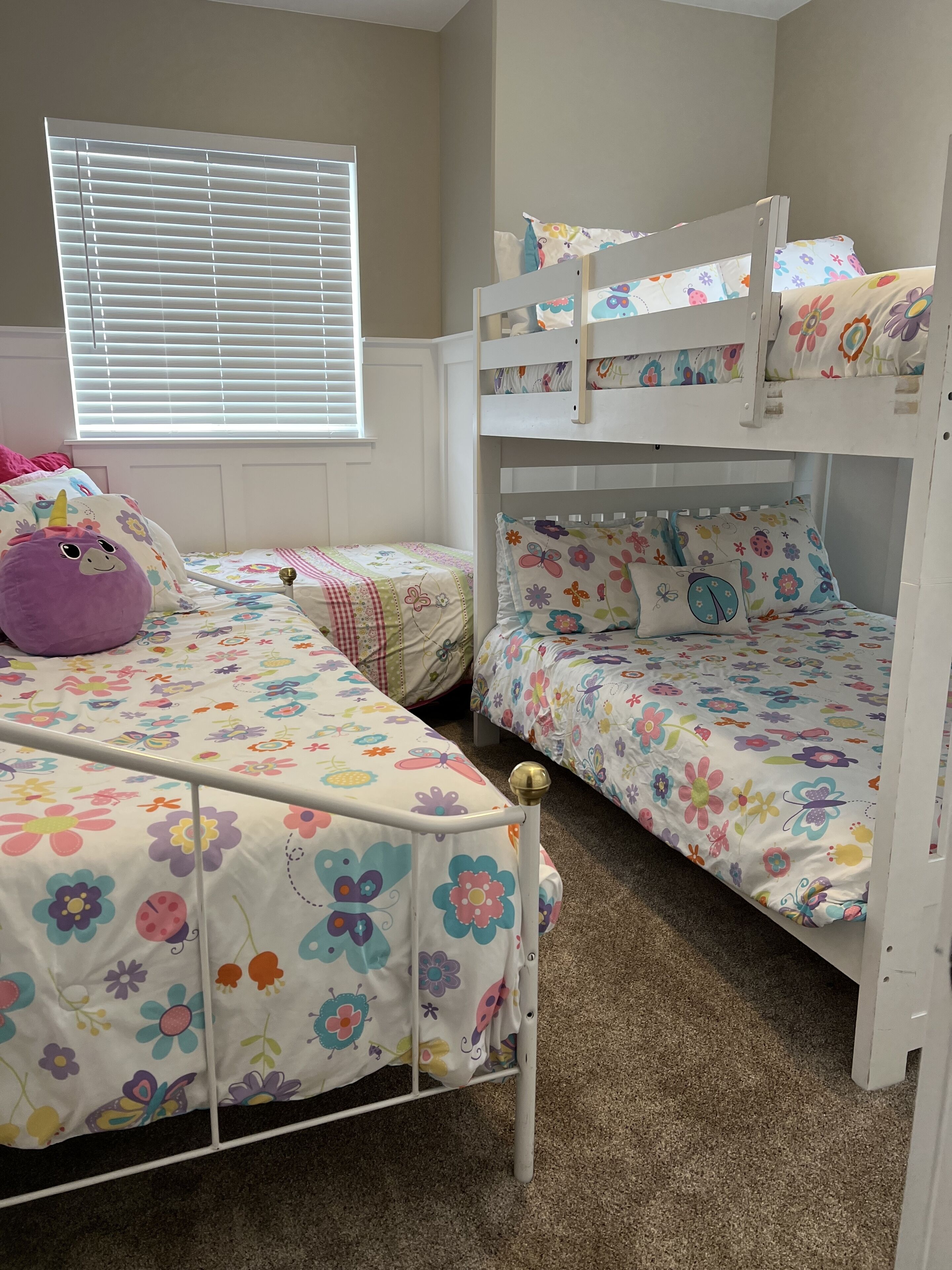 A child's bedroom with bunk beds, floral bedding, a purple unicorn pillow, and a window with blinds.