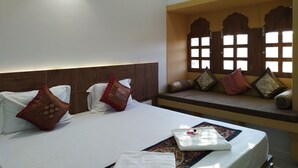 A hotel room with a bed, pillows, a bedside table, and a window with decorative shutters. - Aapni Haveli (Jaisalmer)
