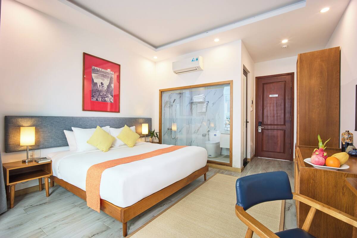 Habitación de hotel con cama, mesitas de noche, un escritorio con fruta, una silla, baño con puerta de vidrio y un cuadro en la pared.