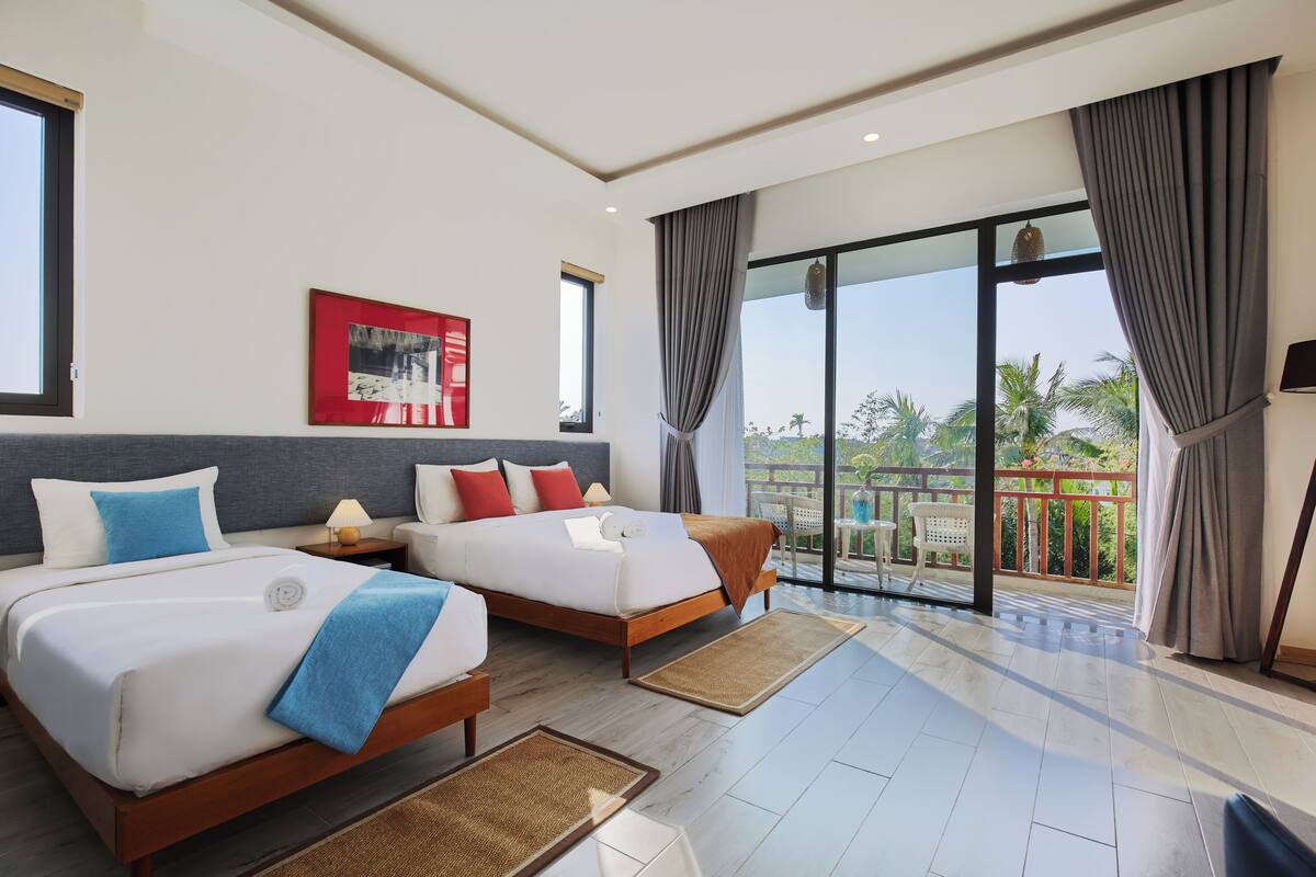 Una habitación de hotel moderna con dos camas, ventanas amplias y un balcón con vistas a palmeras.