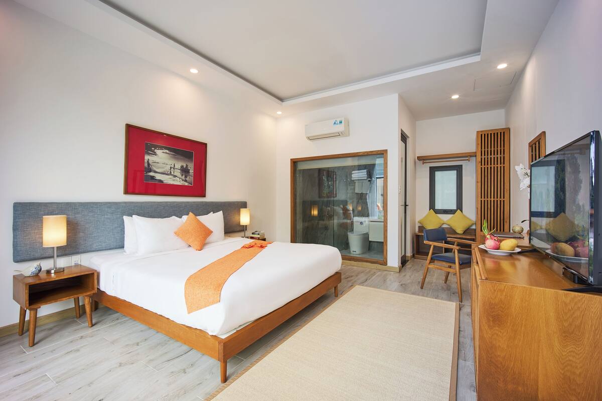 Una habitación de hotel con una cama grande, un escritorio con televisión, una puerta de vidrio que da a un baño y un cuadro en la pared.