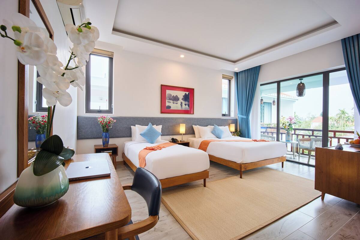 Habitación de hotel con dos camas, un escritorio, una computadora portátil, un florero con flores y un ventanal con vistas.