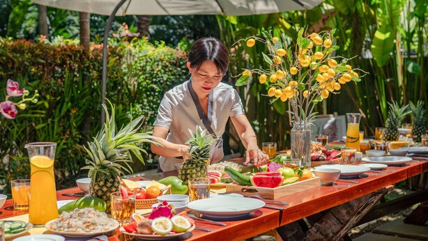 A dining table set outdoors with various fruits, drinks, and a person serving food. - Chieu Ha Village (Da Nang)