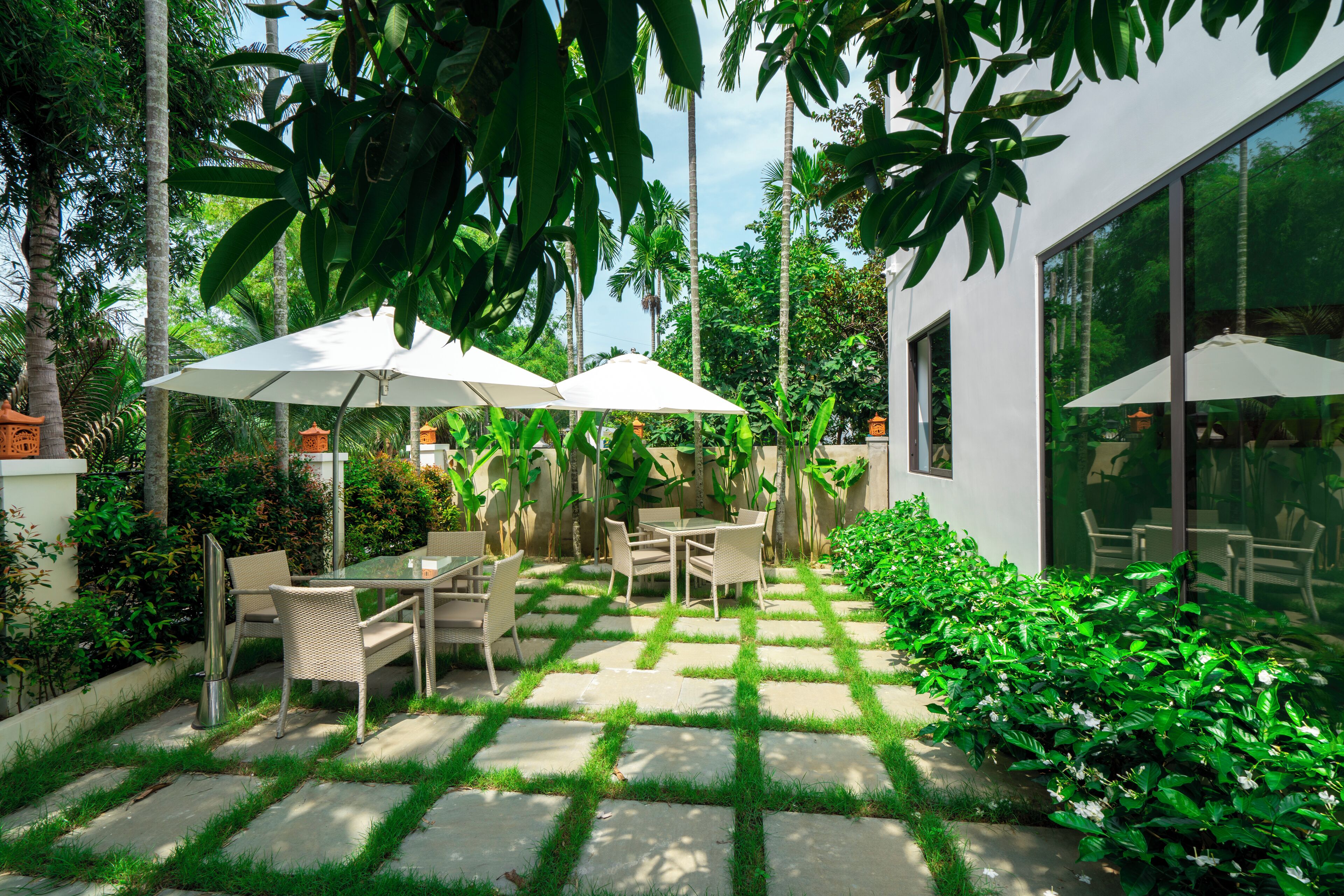 A patio with outdoor furniture and umbrellas, surrounded by lush greenery and a modern building with large windows.