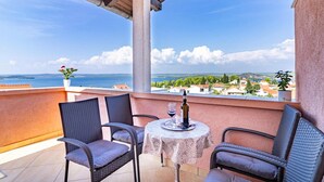 A balcony with wicker chairs, a table with a bottle and glass, and a view of the sea and town.