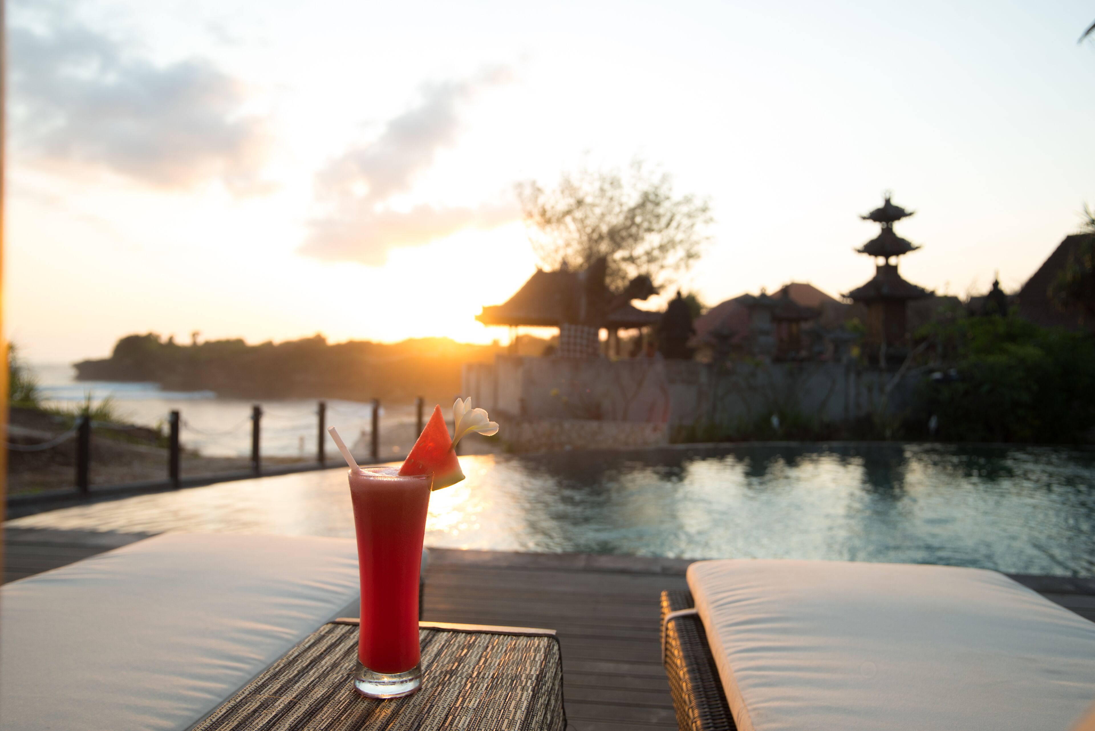 A sunset view from a deck with a drink on a table, overlooking a calm body of water and traditional architecture.