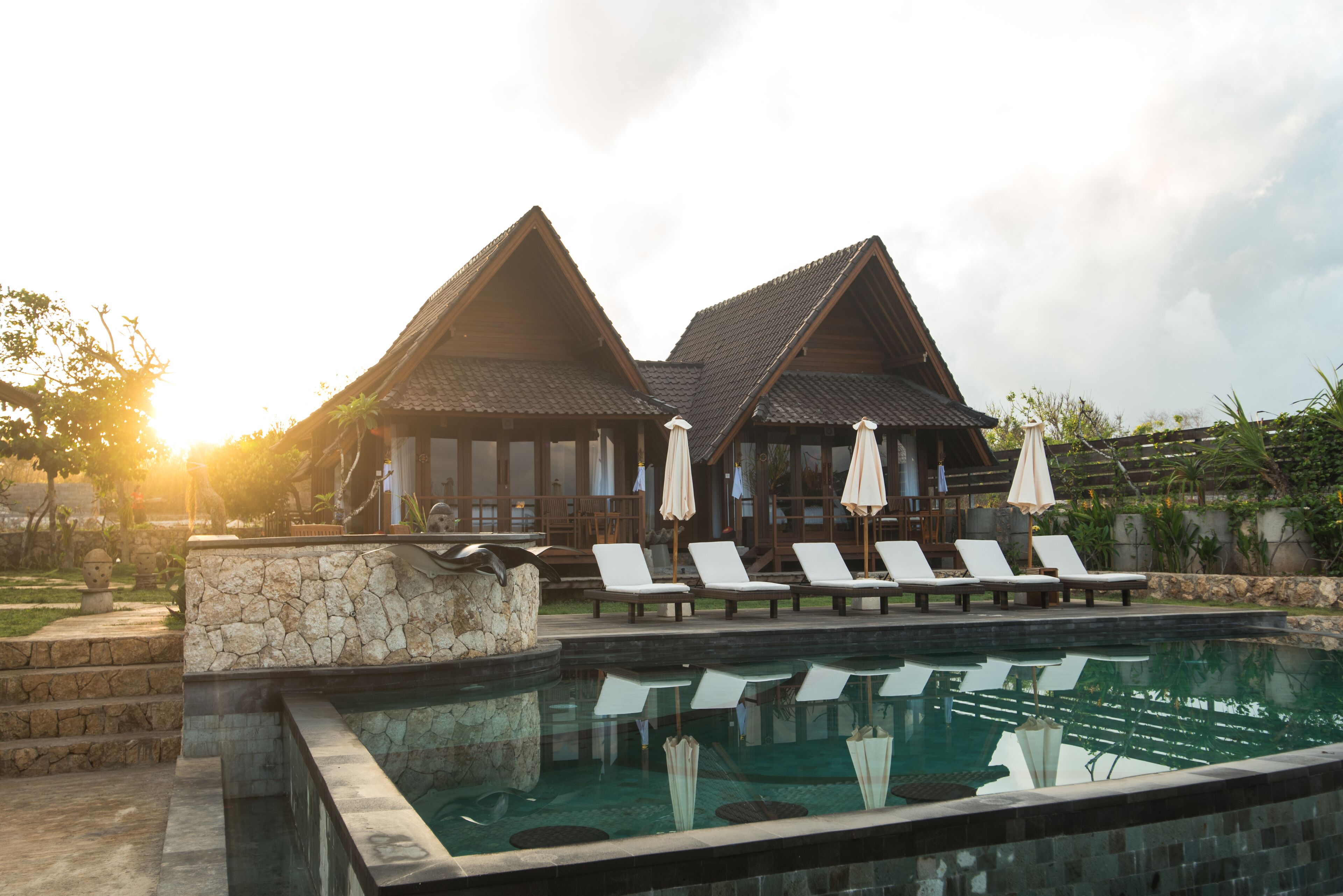 A pool area with lounge chairs and umbrellas, a stone wall, and a building with a tiled roof.