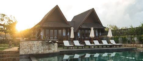 A pool area with lounge chairs and umbrellas, a stone wall, and a building with a tiled roof.
