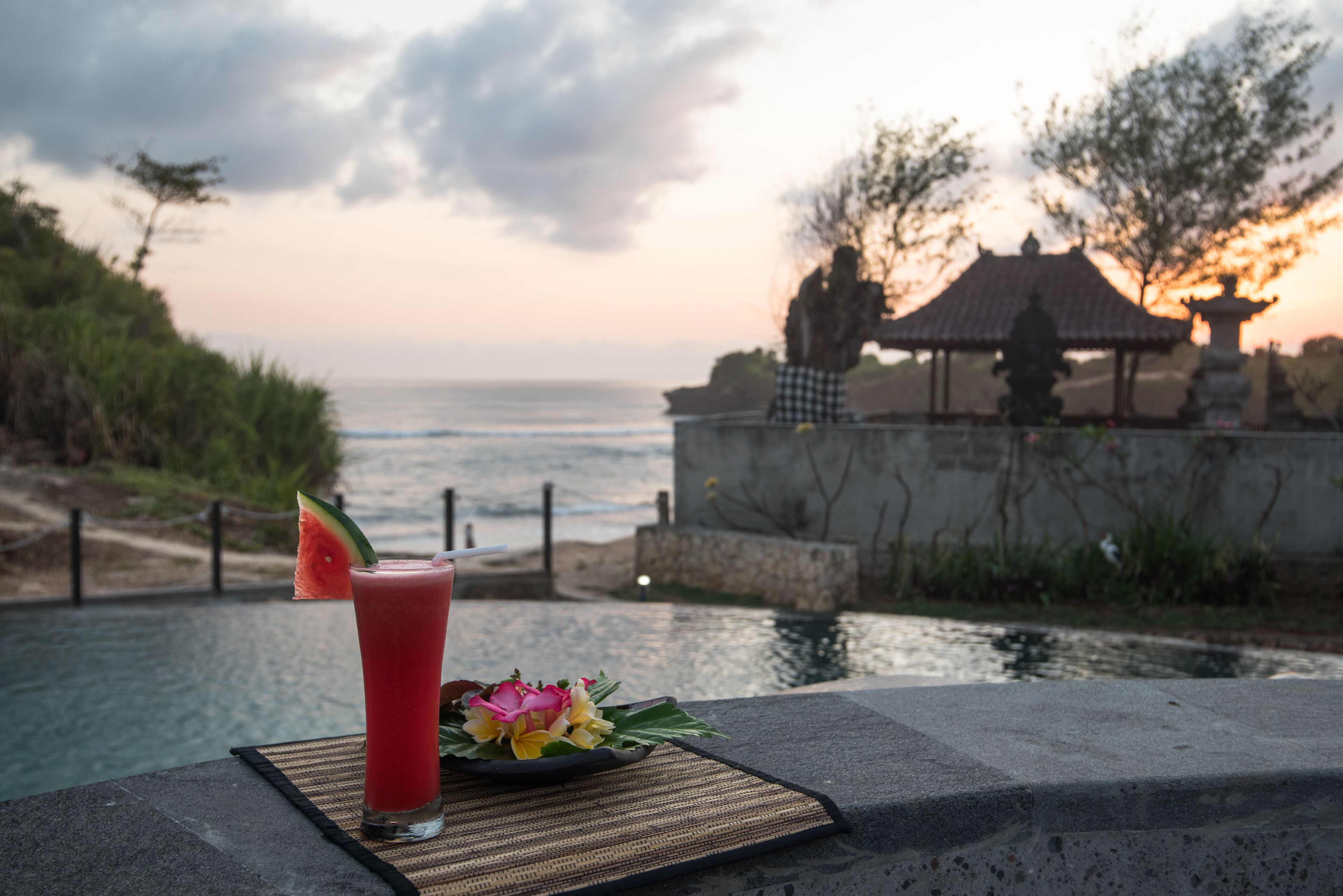 A table with a drink, a flower, and a view of the ocean at sunset.