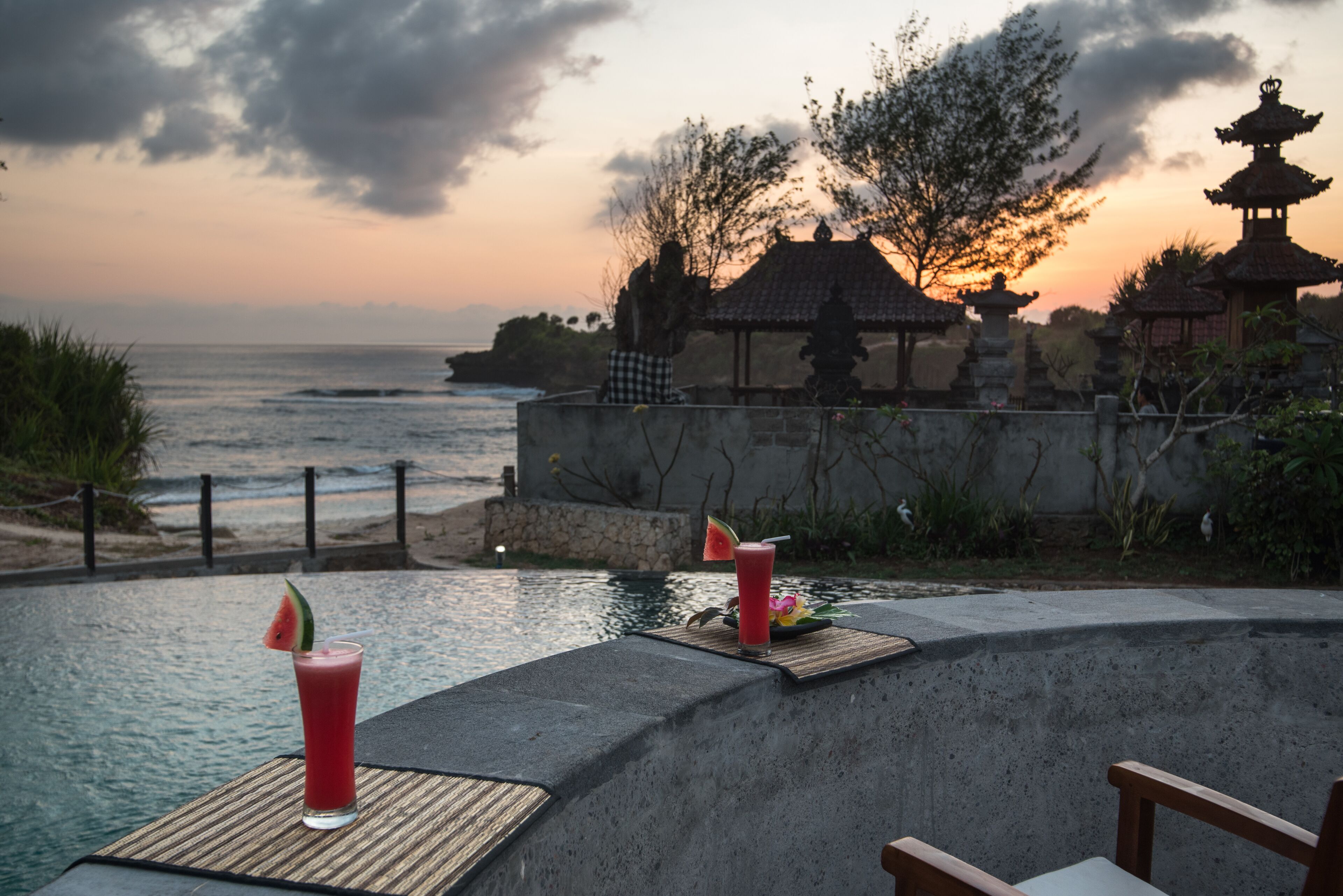 A poolside area with two drinks on a table, overlooking a body of water and traditional architecture in the background.