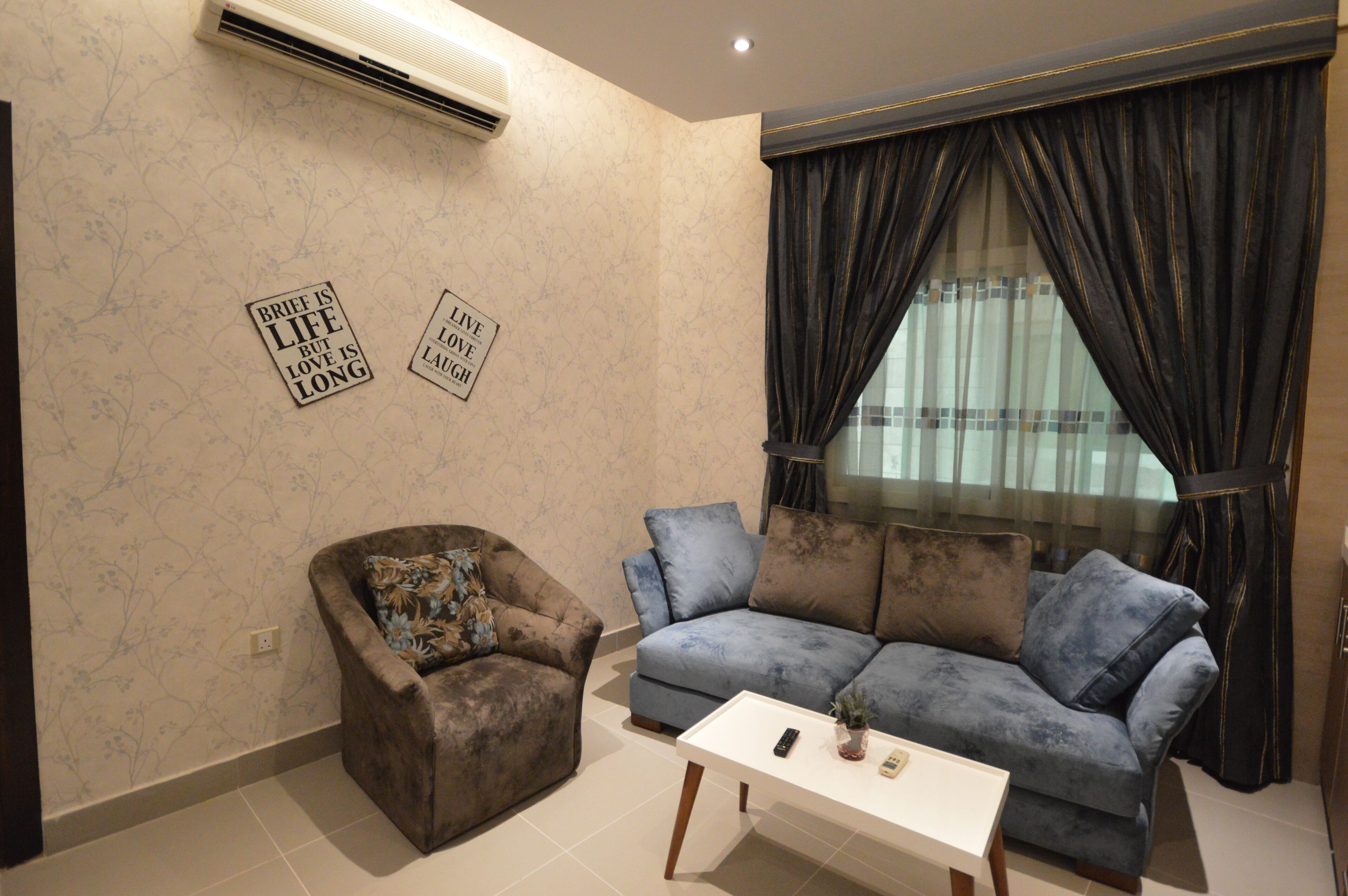 A living room with a brown armchair, a blue sofa, and a white coffee table. There are two wall decorations with text, and a large window with dark curtains.