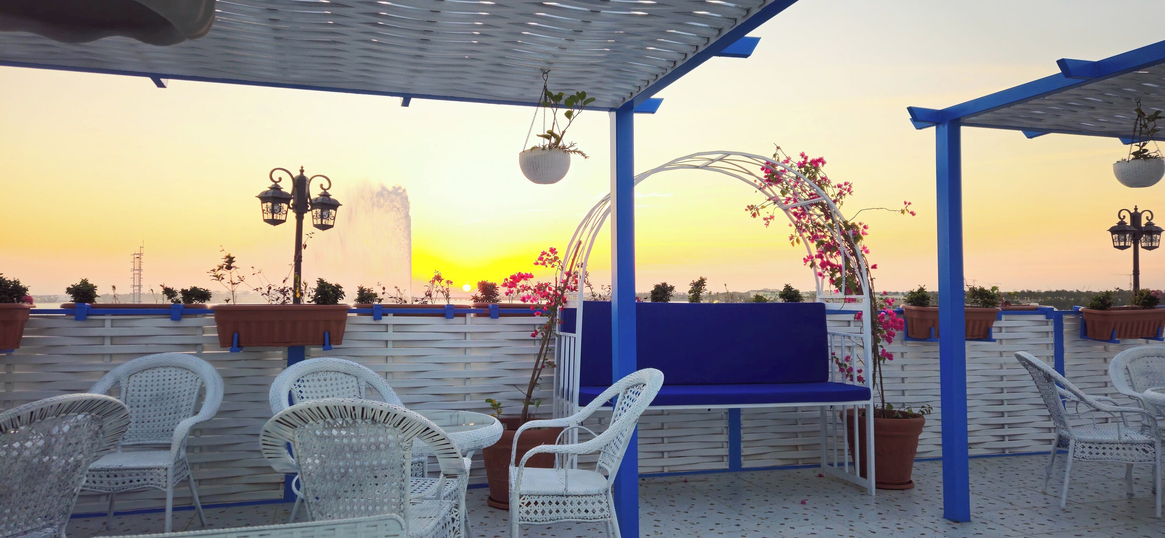 A rooftop terrace with white wicker furniture, a blue bench, and potted plants.