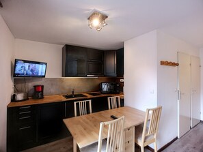 A compact kitchen with a wooden table, two chairs, a TV mounted above the counter, and a coffee maker. - Comfort Ski Soleil - 2P 4 ppl, Ski-in/Ski-out (Les Menuires)
