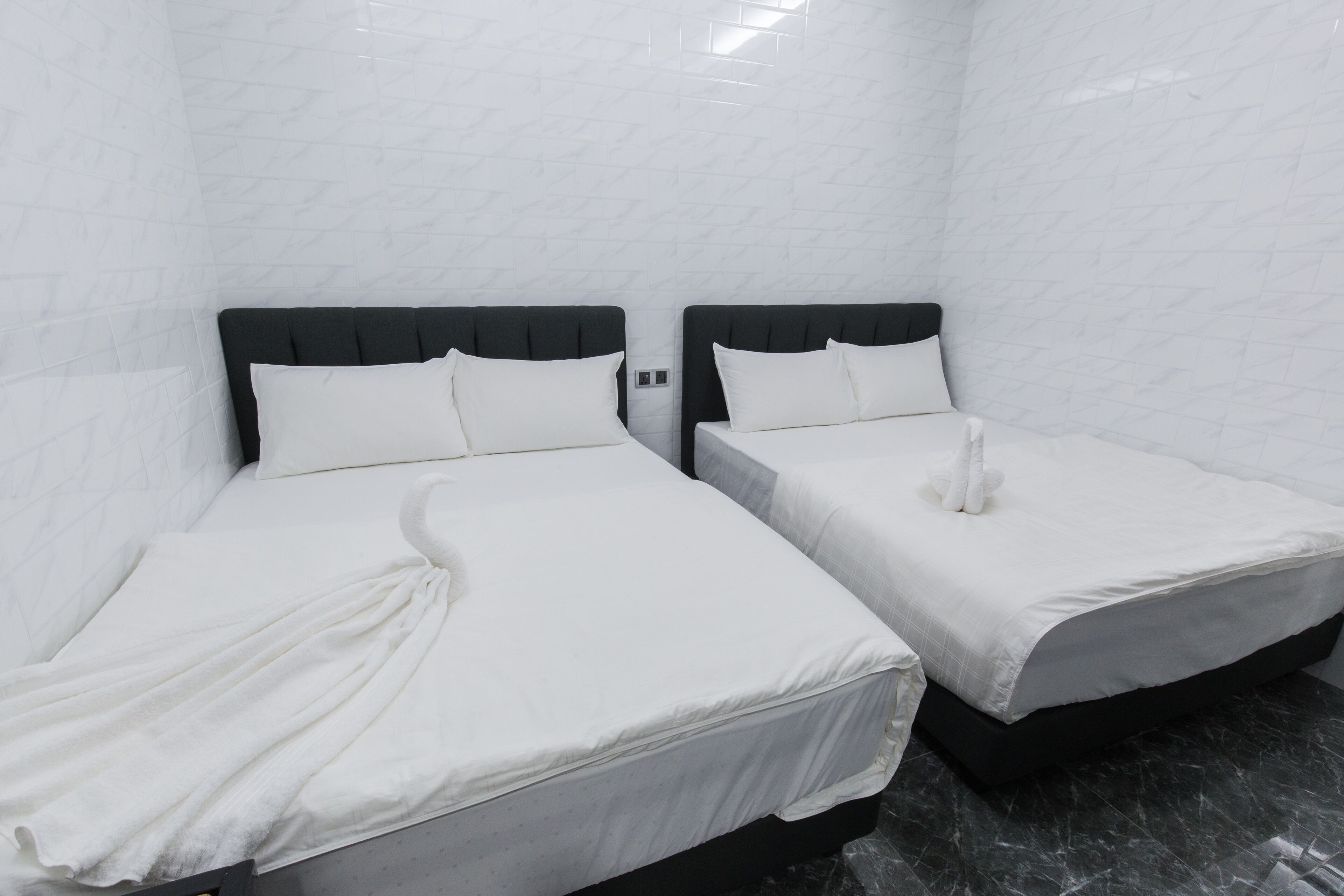 Dos camas individuales con cabeceros negros y ropa de cama blanca en una habitación con paredes de baldosas blancas.