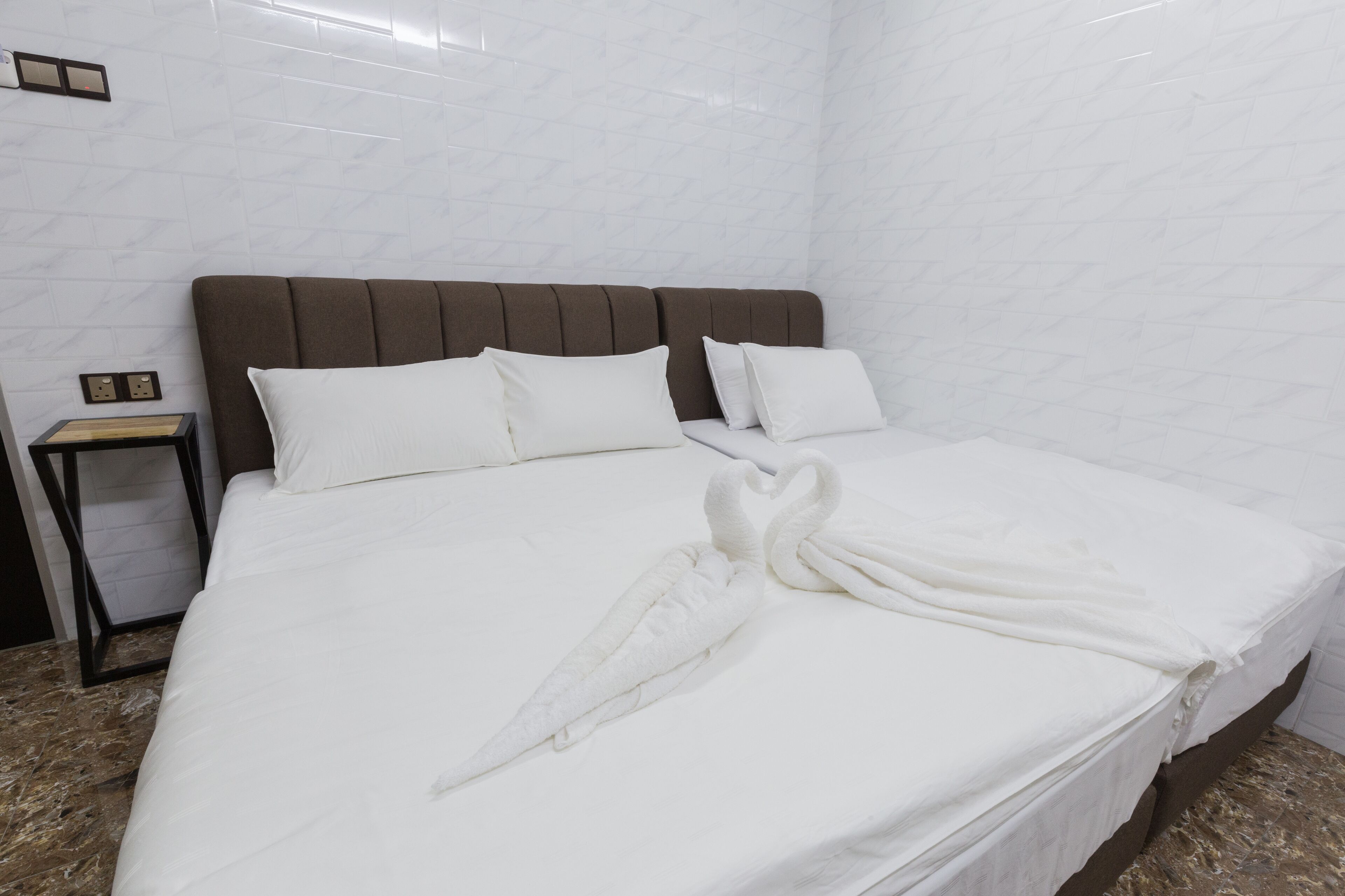 A neatly made bed with white linens and a swan-shaped towel, a brown headboard, and a small bedside table with a lamp.