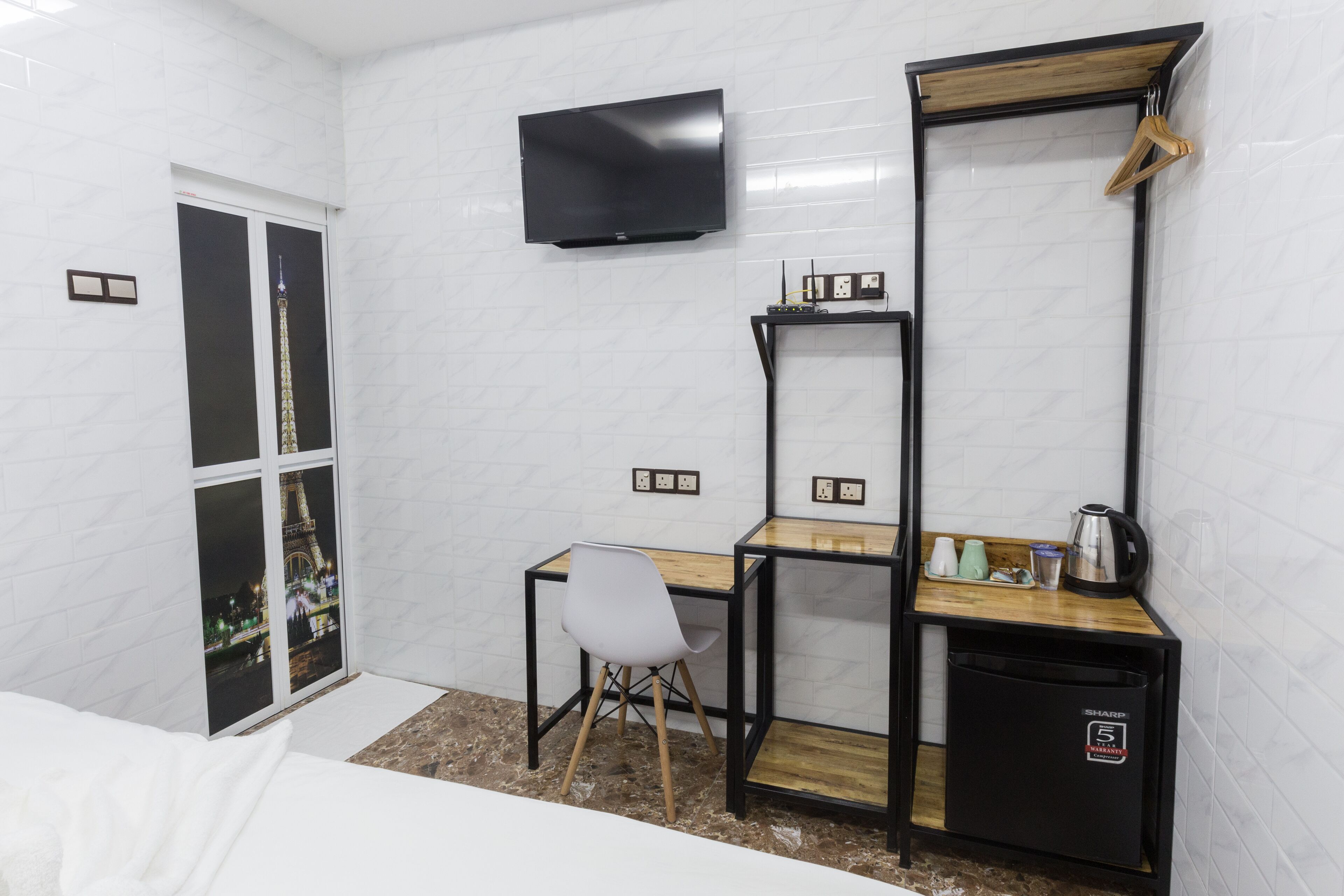 A hotel room with a bed, a desk with a chair, a television mounted on the wall, a mirror, and a small refrigerator.