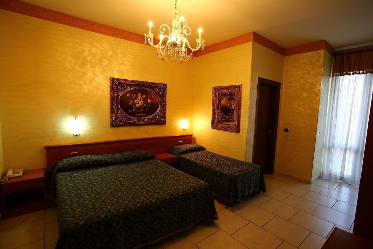 A hotel room with two beds, a chandelier, and two wall-mounted lamps.