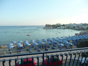 A beach with blue and white striped umbrellas, a few people swimming, and a red car parked near the water. - Hotel Riva del Sole (Porto Cesareo)