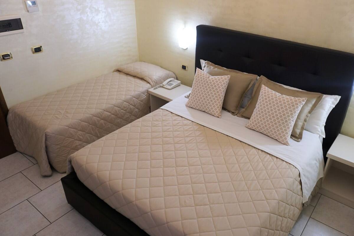 A hotel room with two beds, each with a quilted beige bedspread and matching pillows. There is a bedside table with a telephone and a small shelf.