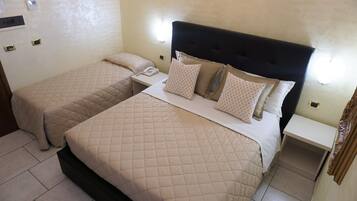 A hotel room with two beds, each with a quilted beige bedspread and matching pillows. There is a bedside table with a telephone and a small shelf.