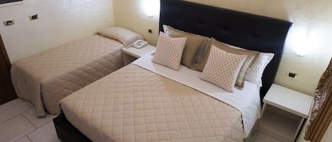 A hotel room with two beds, each with a quilted beige bedspread and matching pillows. There is a bedside table with a telephone and a small shelf.