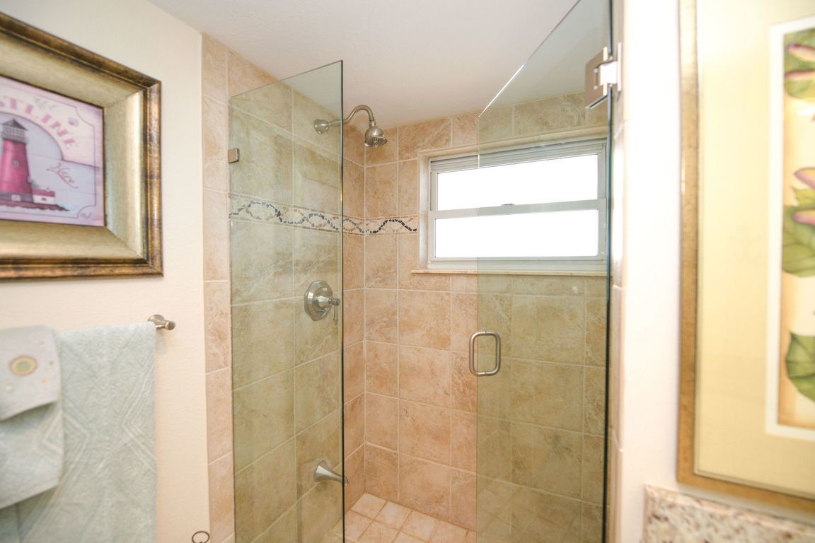 Une douche fermée par des parois de verre, avec des carreaux de couleur beige, une fenêtre et un tableau encadré au mur.