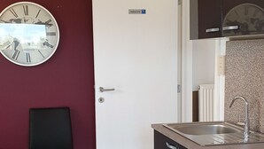 A compact kitchen with a white door, a clock on the wall, and a dining area with a table and chairs. - Housing for expats and companies (Arcelor Mittal, Volvo, ...) (Lochristi)