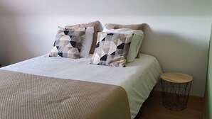 A neatly made bed with patterned pillows, a round wooden side table, and a plain wall. - Housing for expats and companies (Arcelor Mittal, Volvo, ...) (Lochristi)