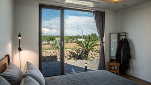 A bedroom with a large sliding glass door leading to a garden, a bed with pillows, a bedside lamp, and a small wooden stool. - This is a modern design space, and you can enjoy outdoor bath facilities (jeju)