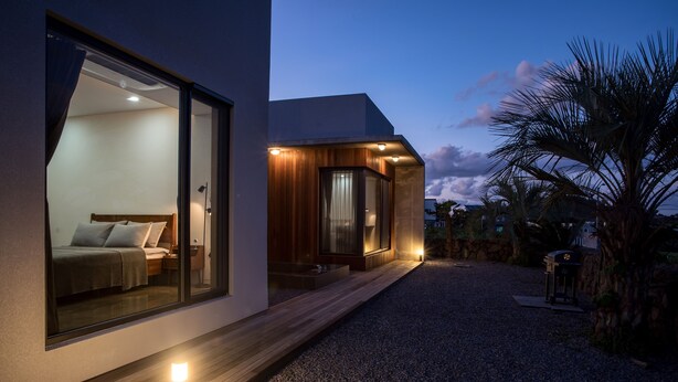 A modern house with a large glass window revealing a bedroom, a wooden exterior, and a gravel driveway. - This is a modern design space, and you can enjoy outdoor bath facilities (jeju)