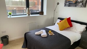 Een netjes opgemaakt bed met een donkere sprei, een wit kussen en een geel kussen. Er staat een klein tafeltje met een glas sinaasappelsap en een boek. Een raam toont uitzicht op een bakstenen muur en een hek.
