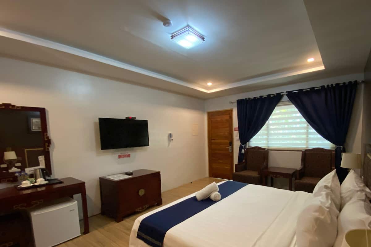 A hotel room with a bed, a desk, a television, a small refrigerator, a window with curtains, and a ceiling light.