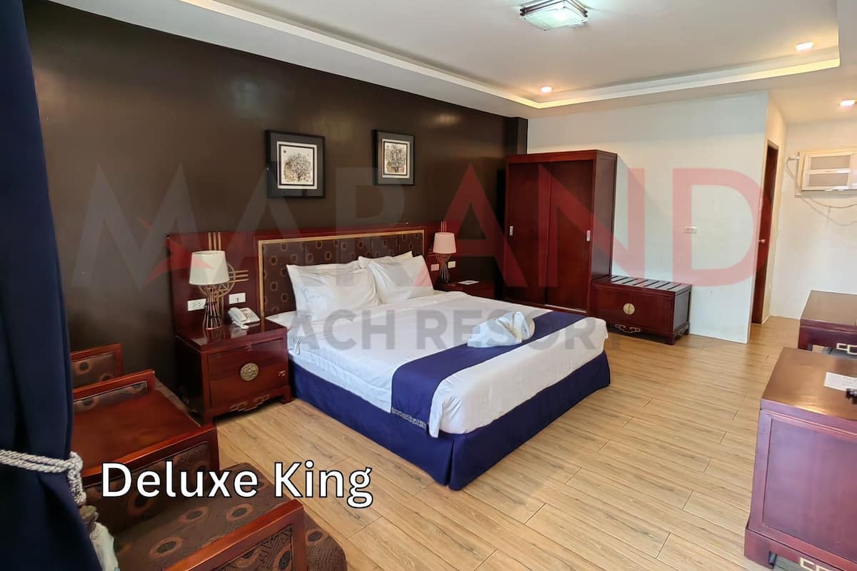A Deluxe King bed in a hotel room with a dark brown wall, two framed pictures, a wooden wardrobe, and a bedside table with a lamp.