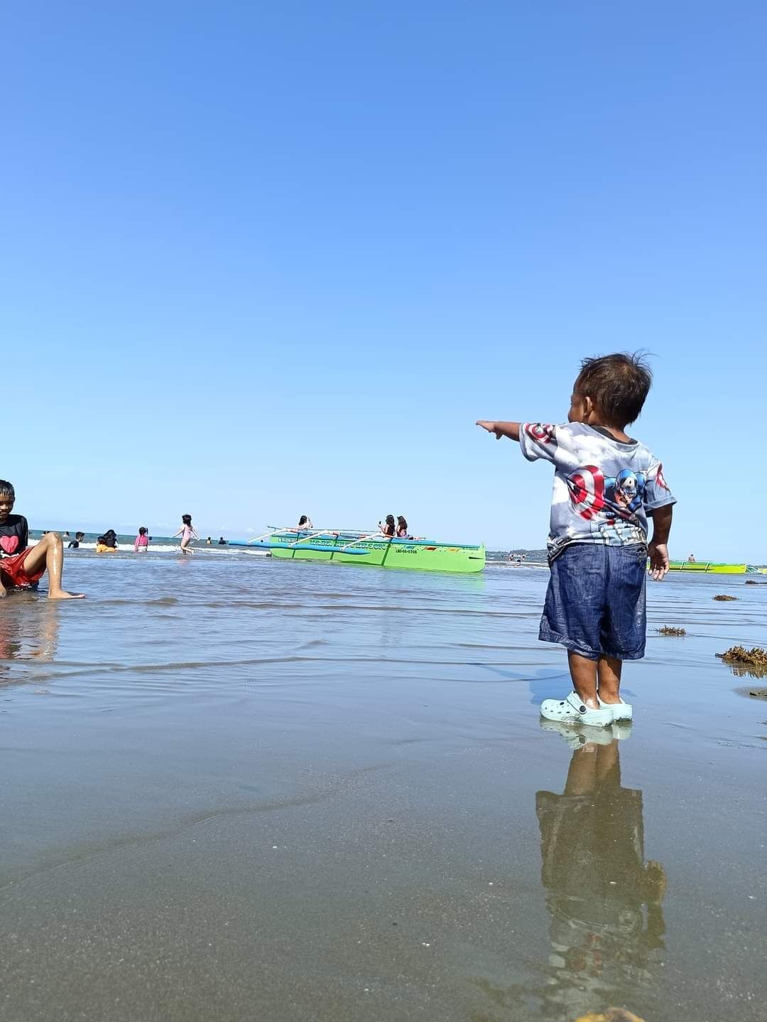 Eine Szene am Strand, ein Kind deutet in Richtung Wasser, ein grünes Boot und Menschen im Hintergrund.