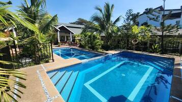 A swimming pool with clear blue water, surrounded by palm trees and a building with a sloped roof.