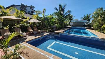 A swimming pool with lounge chairs and umbrellas, surrounded by palm trees and a building.