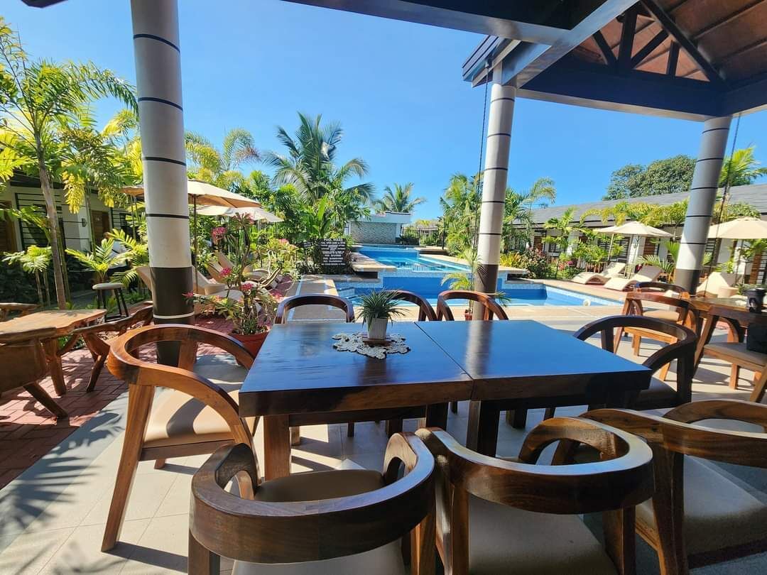 An outdoor dining area with wooden tables and chairs, a pool in the background, and tropical plants.